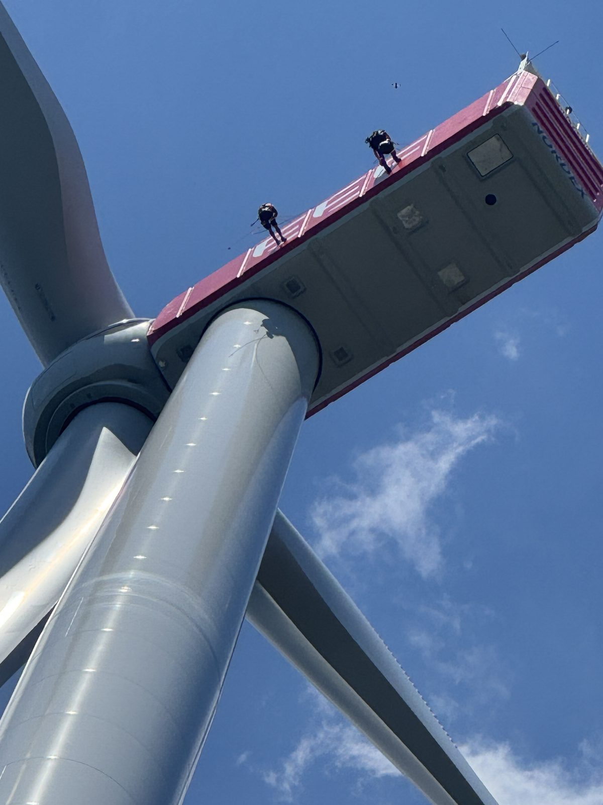 Blick von unten auf die Gondel einer Windkraftanlage mit Industriekletterern auf dem Dach